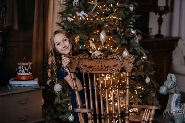 青いドレスのかわいい女の子はクリスマスツリーにポーズをとります 新年の場所に子供 新年の装飾付きの写真スタジオ 子供は移動中です 選択的フォーカス フォーカス解除 — ストック写真