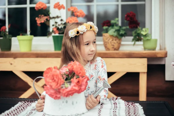 Küçük bir kız taze çiçeklerin arasında bir masada oturuyor. Kafasında çiçek çelengi olan beyaz kazaklı şirin bir kızın portresi. Botanik ve çiçek bakımı kavramı.
