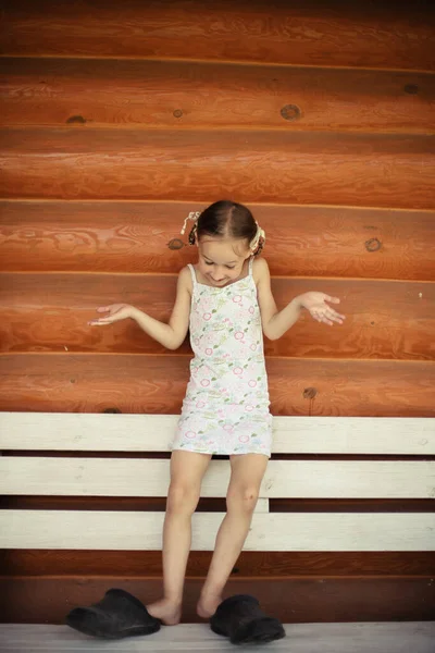 一个穿着白色夏装 笑容可亲的小女孩正在试穿爸爸的拖鞋 一个穿着大拖鞋的孩子在天然原木的背景下 无忧无虑的童年Toning案 — 图库照片