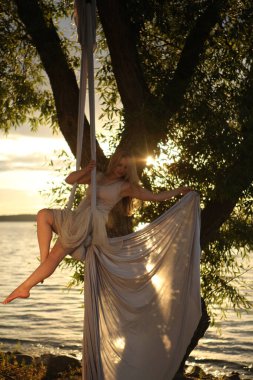 Beyaz elbiseli genç çekici bir kız, açık gökyüzünün arka planında, bir ağacın yanında, doğa sporuna gider. Sağlık ve spor konsepti. Hava jimnastiği.