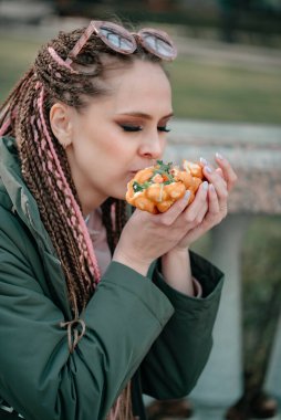 Parlak makyajlı ve afro saçlı genç bir kız bir bankta oturur ve bir sosisli sandviç yer. Seçici odaklı yakın plan fotoğraf. Kafasında güneş gözlüğü olan kız..