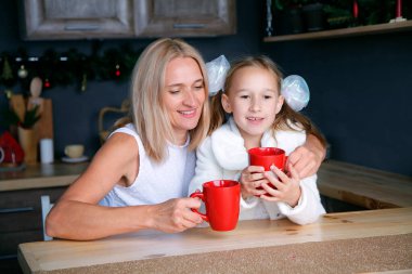 Çekici anne ve kızı yeni yıl mutfağında otururken kırmızı kupalardan sıcak çay içerler. Mutlu Noeller! Tatili bekliyorum..