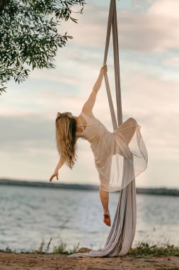 Beyaz elbiseli genç çekici bir kız denizin arka planında, bir ağacın yanında havadan jimnastik yapıyor. Sağlık ve spor konsepti. Deniz kıyısında hava jimnastiği.