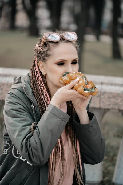 明るいメイクとアフロのピグテールをした若い女の子が公園のベンチに座ってホットドッグを食べています 選択的焦点でクローズアップ写真 頭にサングラスをかけた女の子 — ストック写真