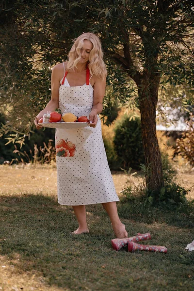 長い白いエプロンをした金髪の幸せそうな女性は 屋外に立っている間 果物や野菜を手にトレイを持っています 夏のピクニック — ストック写真