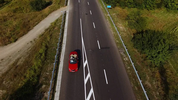 Flying above young couple driving the luxury red convertible car on a countryside road leading through beautiful. Happy man and woman traveling on summer vacation. High quality 4k footage