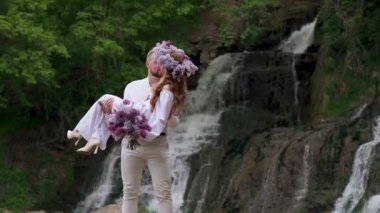 Groom holds bride in his arms near waterfall. Wedding couple. Happy family. Man and woman in love. Lovely groom and bride. Wedding day. Slow motion. High quality FullHD footage