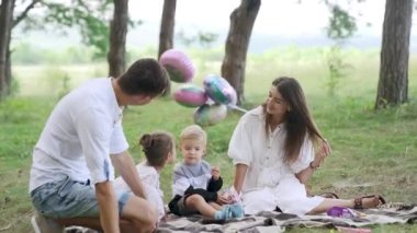 Happy family relax lying picnic blanket. Young people rest on green lawn with two kids summer day. Top view tranquil parents with children on fresh grass near pine trees forest. FullHD footage