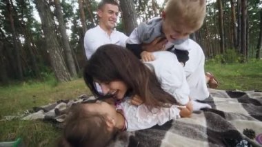 Laughing parents tickling cute kids on picnic blanket wide shot. Parents play with cheerful kids outdoors on summer weekend. Carefree family have fun with two children pampering. FullHD footage