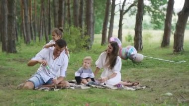 Happy family relax lying picnic blanket. Young people rest on green lawn with two kids summer day. Top view tranquil parents with children on fresh grass near pine trees forest. FullHD footage