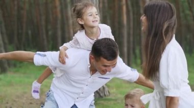 Beautiful girl sitting between smiling parents in wheat field and hugging them. Loving family having picnic in pine forest. Parents with two kids. High quality FullHD footage