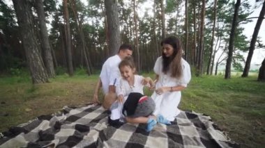 Laughing parents tickling cute kids on picnic blanket wide shot. Parents play with cheerful kids outdoors on summer weekend. Carefree family have fun with two children pampering. FullHD footage
