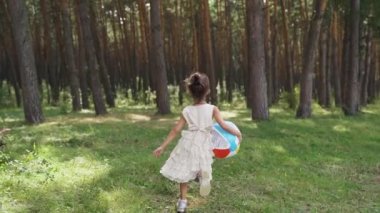 Little child girl walks through park with bright colored ball. Concept children dream game. Daughter kid plays ball field. child runs with ball along road sunset. Slow motion. FullHD footage