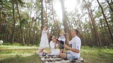 Happy family including mother, father, and two kids are blowing soap bubbles and enjoying the summer holidays in the park. Smiling parents and children are spending leisure time together. FullHD