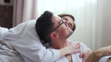 Young happy couple smiling having fun, resting lying on bed, loving husband and wife embracing kissing spending time together at home in bedroom, enjoying comfortable relaxation in the morning. FullHD