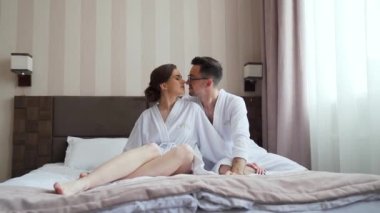 Young couple in bathrobe enjoying together time in wellness hotel room, lying in the bed and hugging. High quality FullHD footage