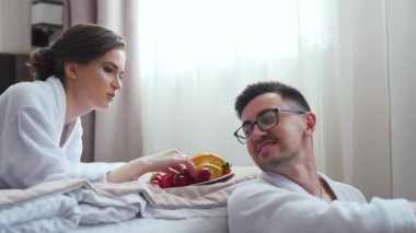 Happy lovely couple dressed in bathrobes lying together on bed, eating fruits and talking. Newlywed in hotel room. High quality FullHD footage