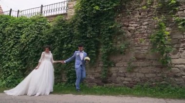 Peçeli dar gelinlik giymiş gelin, nişanlısının damat elini turuncu tuğlalı duvara karşı tutmasını takip ediyor. Yüksek kaliteli FullHD görüntüler