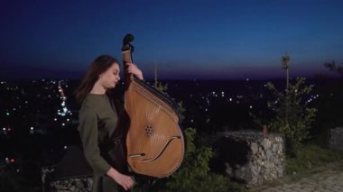 Şaşkın bakışlı bir kız, gece şehir arka planında bandura enstrümanı çalıyor. Yüksek kaliteli FullHD görüntüler