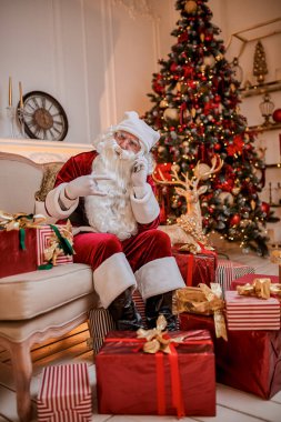 Noel Baba kanepede oturuyor ve cep telefonuyla şöminenin yanında ve Noel ağacında hediyelerle konuşuyor. Yeni yıl ve Mutlu Noeller, Mutlu Noeller konsepti