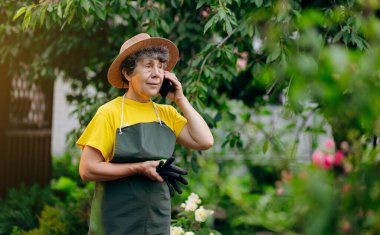 Bahçesinde çalışan şapkalı bir bahçıvan kadın telefonda konuşuyor. Bahçe işleri, çiçek ve bitkilerle ilgilenmek ve büyümek..
