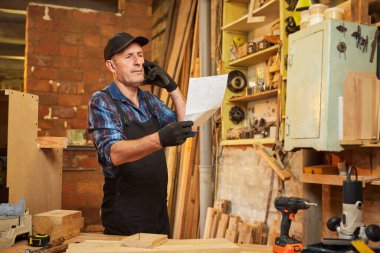 Üst düzey bir marangozun portresi mobilya yapma planlarına bakıyor ve marangozluk atölyesindeki bir müşteriyle telefonda konuşuyor.
