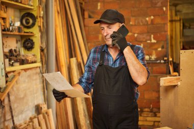 Üst düzey bir marangozun portresi mobilya yapma planlarına bakıyor ve marangozluk atölyesindeki bir müşteriyle telefonda konuşuyor.