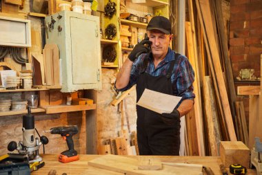 Üst düzey bir marangozun portresi mobilya yapma planlarına bakıyor ve marangozluk atölyesindeki bir müşteriyle telefonda konuşuyor.