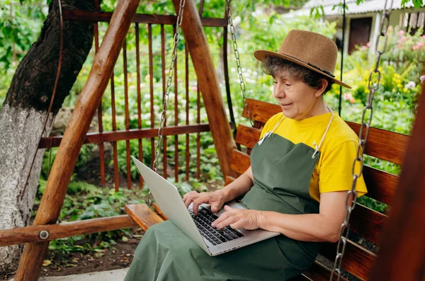 戴着帽子的资深园艺师正在笔记本电脑上观看着什么 并在户外的院子里打了一个视频电话 — 图库照片
