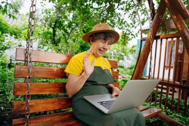 Şapkalı bahçıvan kadın dizüstü bilgisayarından bir şey izliyor ve dışarıdayken bahçede video görüşmesi yapıyor..