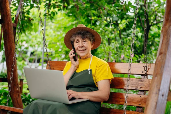 戴着帽子的资深园艺师正在笔记本电脑上观看着什么 并在户外的院子里打了一个视频电话 — 图库照片