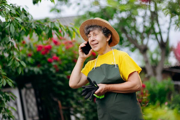 一位戴着帽子的资深园丁在自家院子里干活 还在电话里说着话 种植和照料花卉和植物的概念 — 图库照片