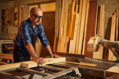 Elektrikli testere ile ahşap kesme masası. Kıdemli profesyonel marangoz üniformalı kereste fabrikasında ahşap tahta kesiyor. Testere makinesi