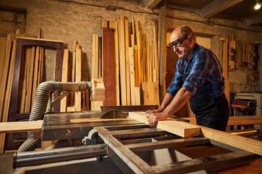 Elektrikli testere ile ahşap kesme masası. Kıdemli profesyonel marangoz üniformalı kereste fabrikasında ahşap tahta kesiyor. Testere makinesi