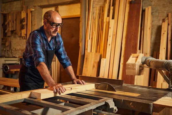 Elektrikli testere ile ahşap kesme masası. Kıdemli profesyonel marangoz üniformalı kereste fabrikasında ahşap tahta kesiyor. Testere makinesi