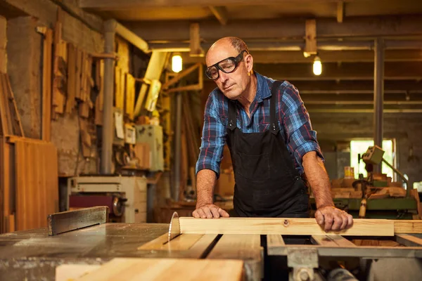 Elektrikli testere ile ahşap kesme masası. Kıdemli profesyonel marangoz üniformalı kereste fabrikasında ahşap tahta kesiyor. Testere makinesi