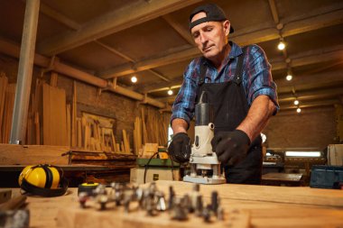 Kıdemli profesyonel marangoz üniformalı marangozluk atölyesinde elle değirmen makinesinde çalışıyor.