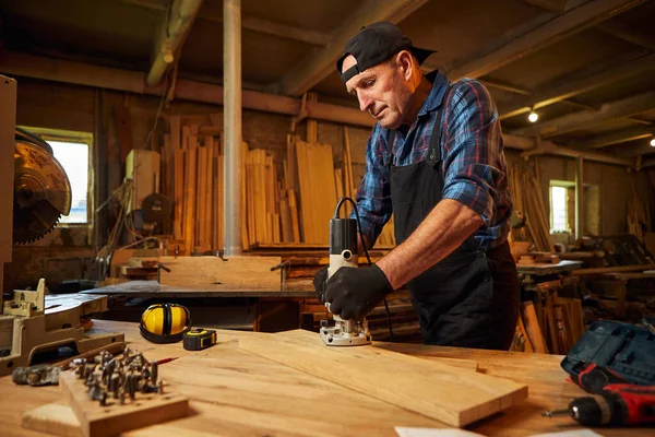 Kıdemli profesyonel marangoz üniformalı marangozluk atölyesinde elle değirmen makinesinde çalışıyor.