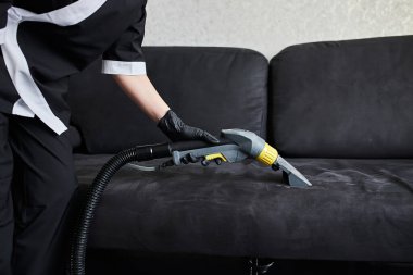 Temizlik şirketi çalışanı, profesyonel malzemelerle mobilyaların tozunu alıyor. Kadın hizmetçi kol temizleme koltuğu elektrikli süpürgeyle kapatılıyor.