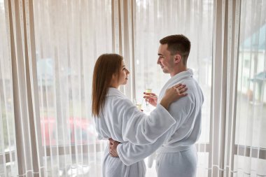 Güzellik, kaplıca, sağlıklı yaşam tarzı konsepti. Lüks otel odasında dinlenen bornozlu genç ve güzel bir çift. Oğlan ve kız rahat bir yerde dinleniyorlar ve şarap içiyorlar. Sağlık teması