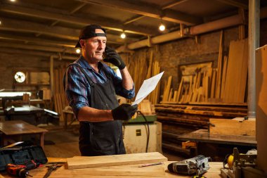 Üst düzey bir marangozun portresi planlara bakıyor ve marangozluk atölyesindeki bir müşteriyle telefonda konuşuyor.