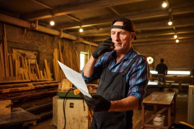 Üst düzey bir marangozun portresi planlara bakıyor ve marangozluk atölyesindeki bir müşteriyle telefonda konuşuyor.