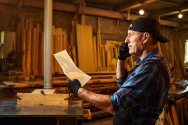 Üst düzey bir marangozun portresi planlara bakıyor ve marangozluk atölyesindeki bir müşteriyle telefonda konuşuyor.