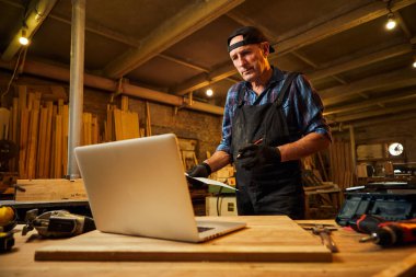 Marangoz, marangozluk atölyesinde dizüstü bilgisayarın yanında duran planlara bakıyor.