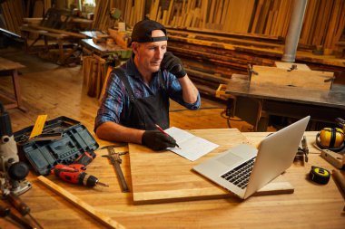 Marangoz bilgisayarda çalışır ve atölyedeki müşteriyle telefonda konuşur.