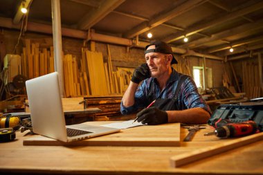 Kıdemli marangoz bilgisayarda çalışır ve atölyedeki müşteriyle telefonda konuşur.