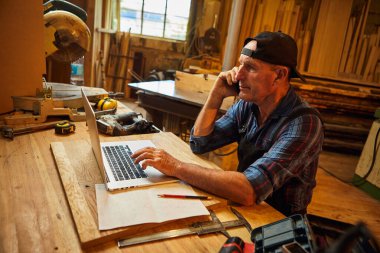 Marangoz bilgisayarda çalışır ve atölyedeki müşteriyle telefonda konuşur.