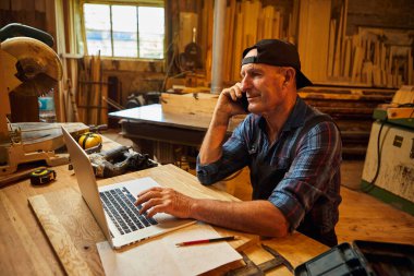 Marangoz bilgisayarda çalışır ve atölyedeki müşteriyle telefonda konuşur.