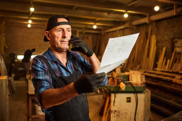 Üst düzey bir marangozun portresi planlara bakıyor ve marangozluk atölyesindeki bir müşteriyle telefonda konuşuyor.