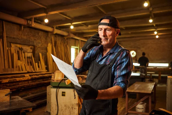 Üst düzey bir marangozun portresi planlara bakıyor ve marangozluk atölyesindeki bir müşteriyle telefonda konuşuyor.